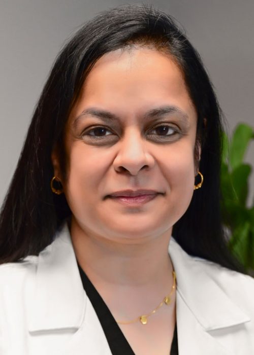 Professional headshot of a woman with long black hair wearing a white lab coat and gold jewelry, smiling slightly in a medical office setting.